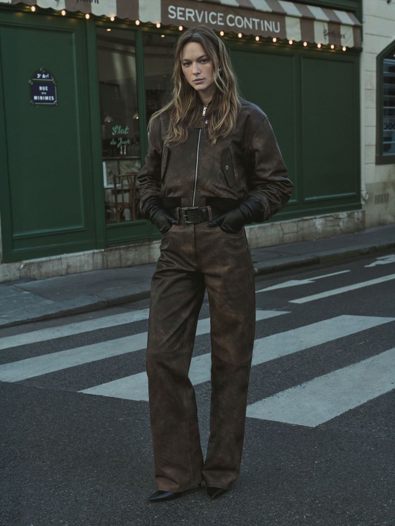 Model in brown leather jacket and matching pants standing on a city street in front of a green storefront.