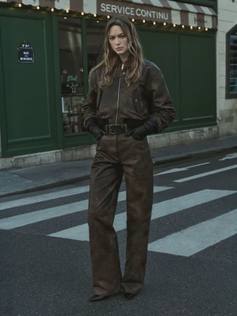 Model in brown leather jacket and matching pants standing on a city street in front of a green storefront.