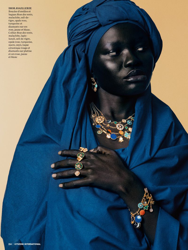 Close-up of a dark-skinned woman wearing a blue headscarf, dress, and jewelry including a bracelet, necklace, earrings, and rings.