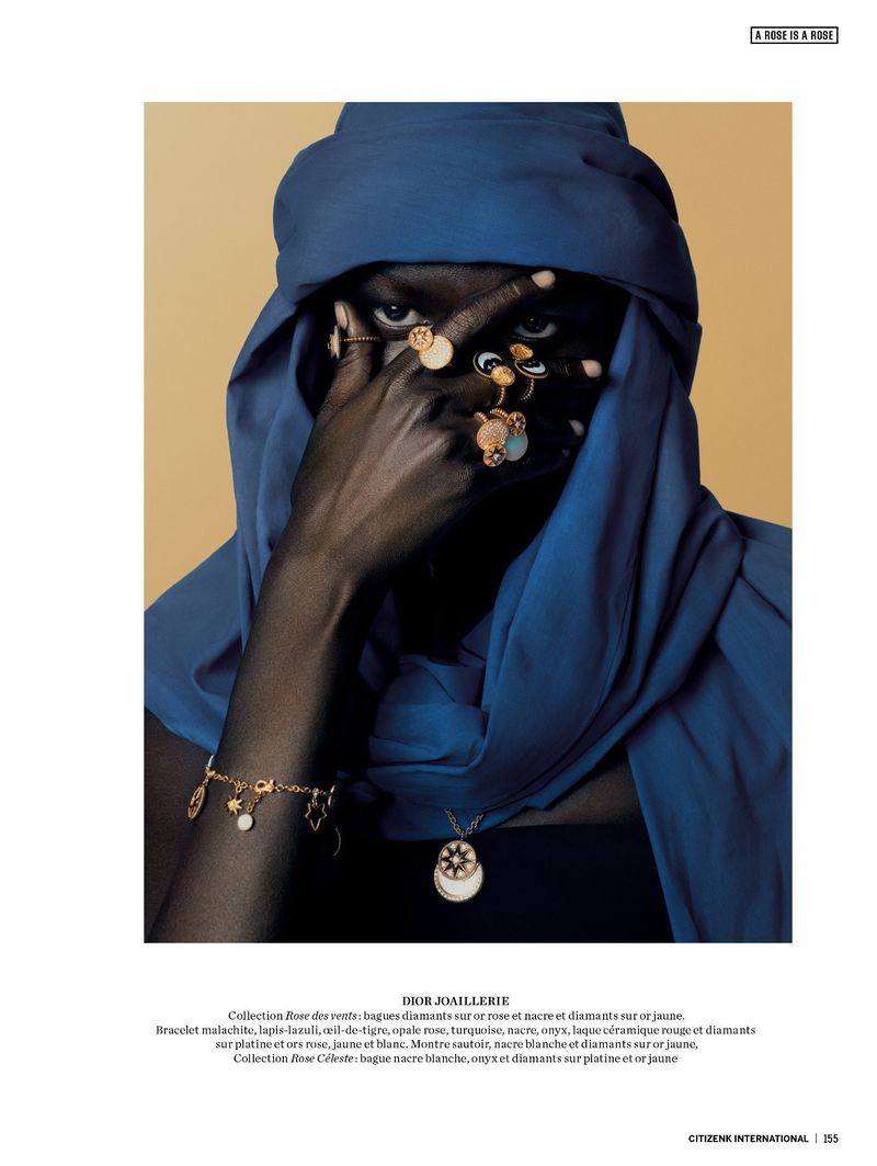 A dark-skinned model wearing a blue turban and jewelry on their face, hands, and neck, standing in front of a tan background.