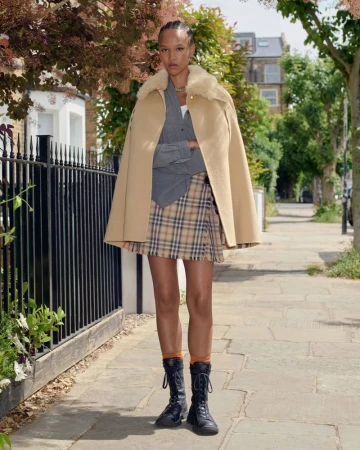 Model standing on a London sidewalk wearing a beige cape, plaid skirt, and black boots against a residential street background.