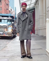 A man wearing a grey trench coat and a red turban stands on a city sidewalk next to a large truck.
