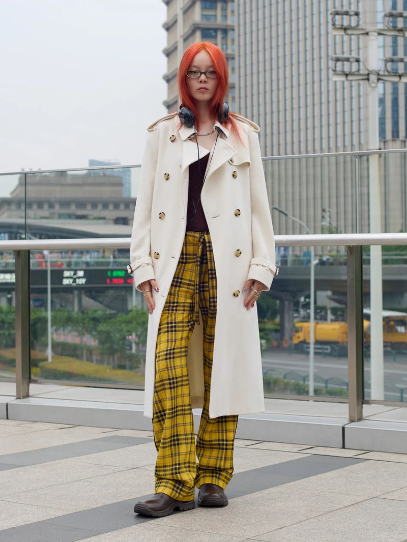Model with vibrant orange hair wearing a cream trench coat and yellow plaid trousers outdoors in an urban setting