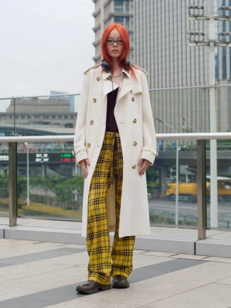 Model with vibrant orange hair wearing a cream trench coat and yellow plaid trousers outdoors in an urban setting