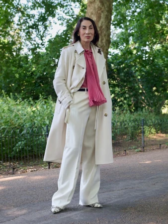 Elderly woman standing on a paved path wearing an off-white trench coat over white trousers and a pleated pink top.