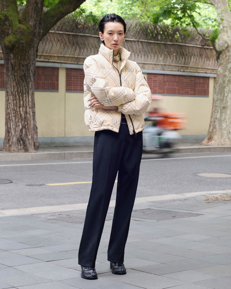 Model standing outdoors wearing a cream plaid puffer jacket and dark trousers on a city street background.