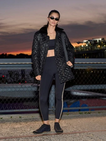 Model wearing black sunglasses and a quilted jacket standing outdoors against a twilight sky and bridge railing background.