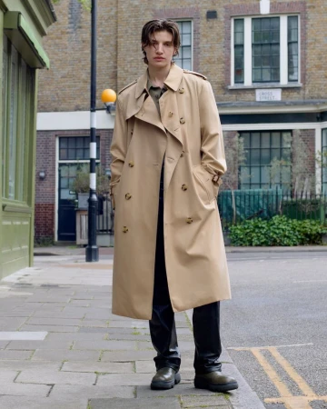 Model standing on a city street wearing a long tan trench coat over dark trousers for Burberry