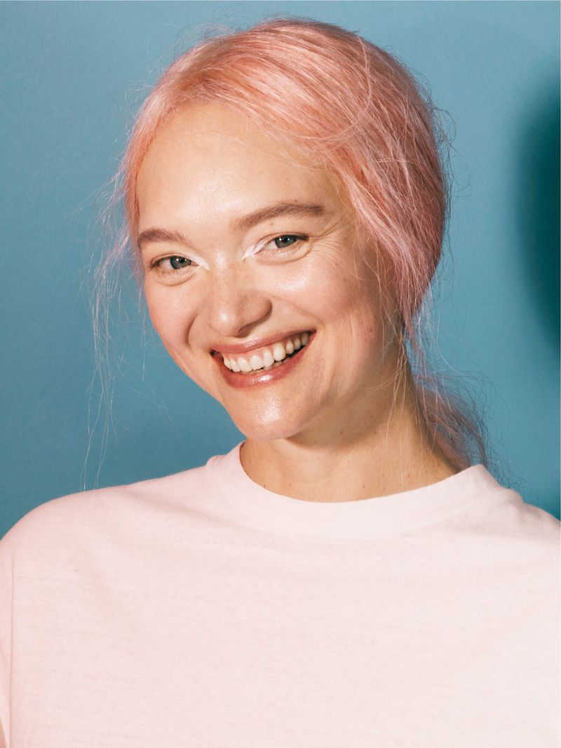Portrait of a smiling woman with pink hair and a light pink t-shirt against a blue background
