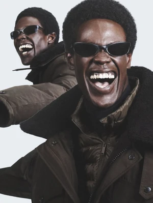 Two black men smiling in winter parkas and sunglasses against a white background