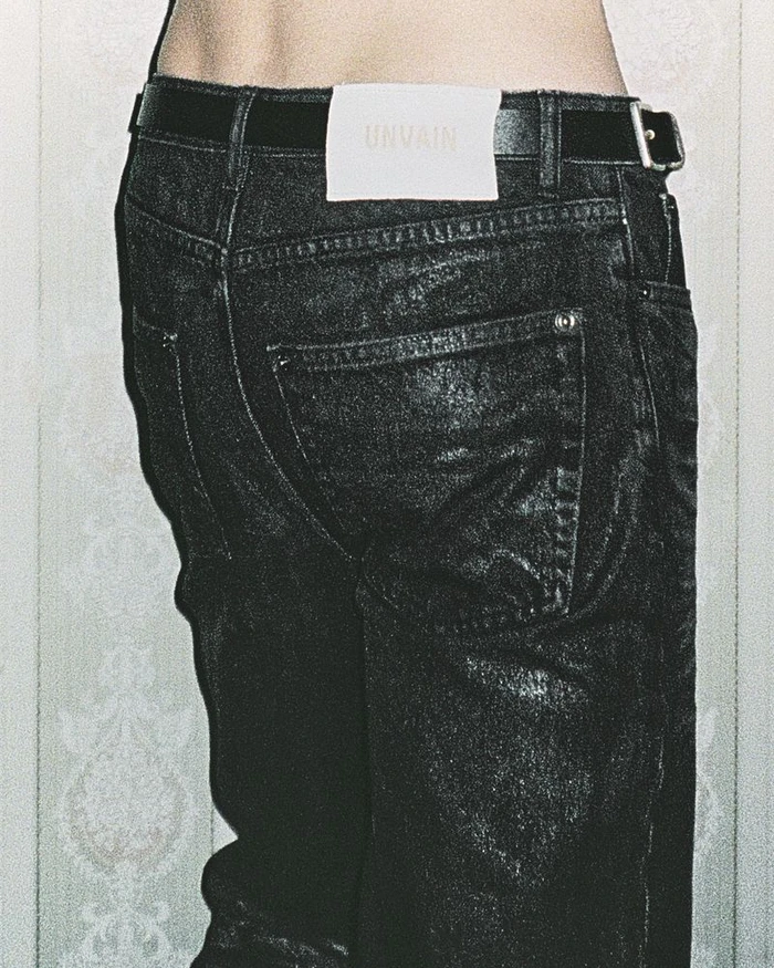 Close-up of black denim jeans with a white branded leather patch and black belt against a textured wallpaper background.