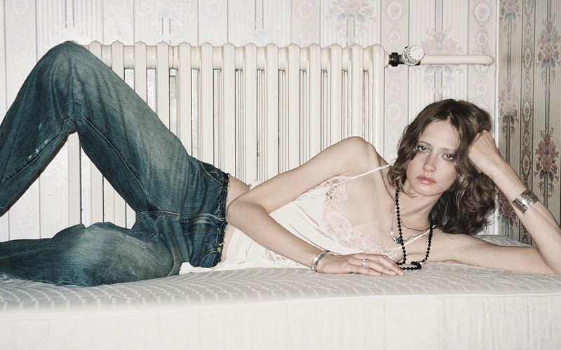 Model lying on a mattress wearing blue jeans and a white camisole against a radiator and floral wallpaper