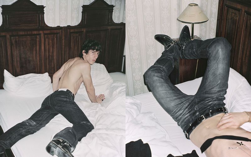 Shirtless young man in dark jeans reclining on a bed in a hotel room with dark wooden headboard and patterned wallpaper.