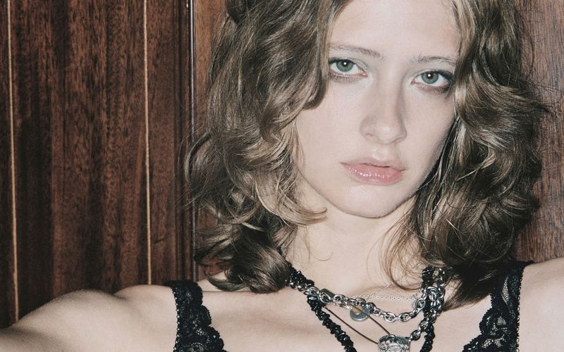 Close-up portrait of a woman with layered brown hair wearing a black lace top and silver chain necklaces against wood paneling
