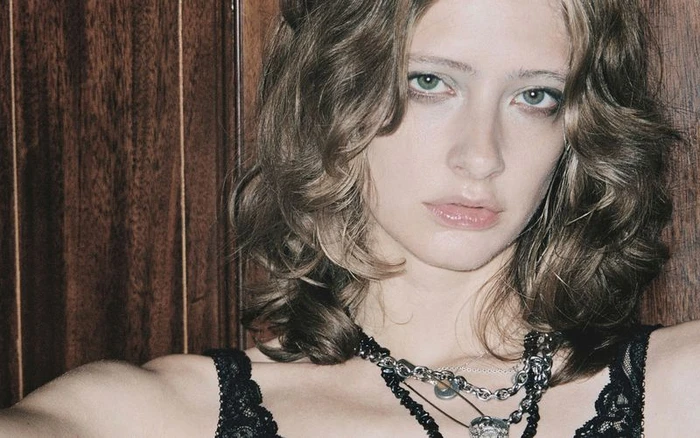 Close-up portrait of a woman with layered brown hair wearing a black lace top and silver chain necklaces against wood paneling