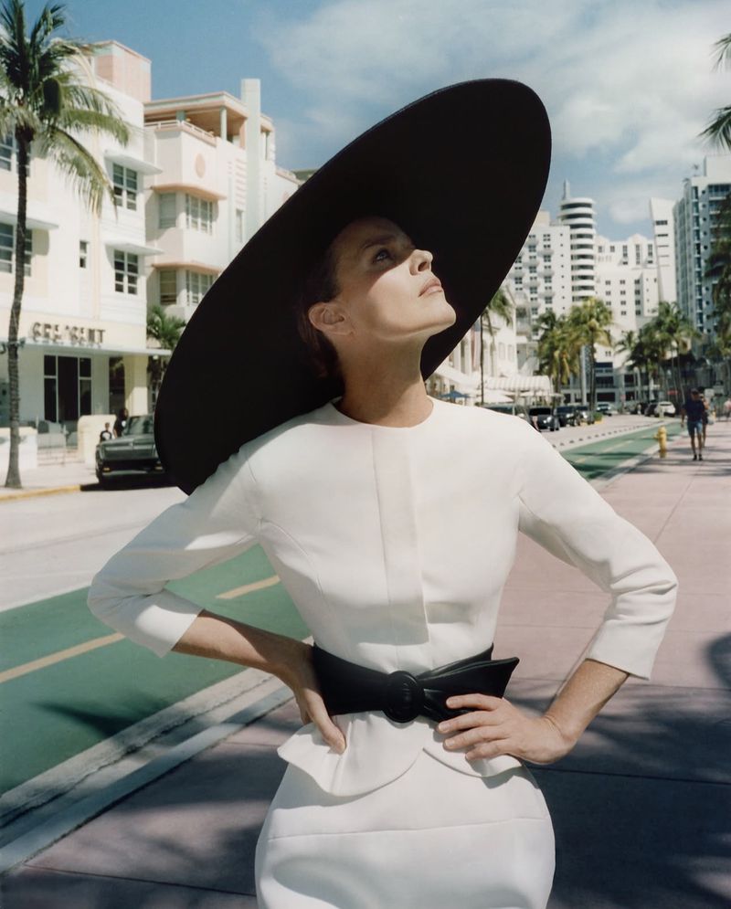 Model wearing a white dress and large black wide-brim hat standing on a sunny urban street
