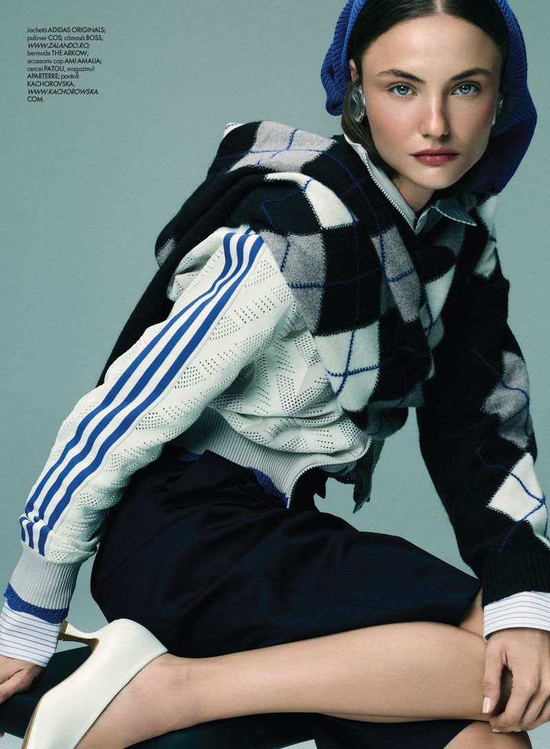 Model Paula Cioltean wearing argyle knitwear and a blue hood against a solid backdrop for Elle Romania.