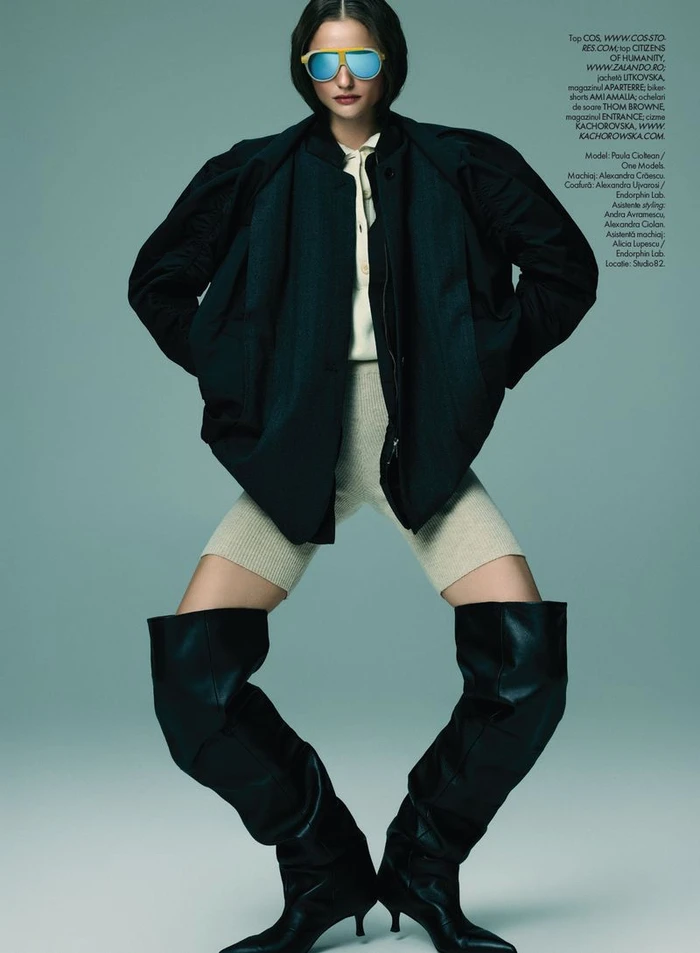 Model wearing blue mirrored sunglasses, dark jacket, cream top, knit shorts, and tall black boots against a plain grey backdrop.