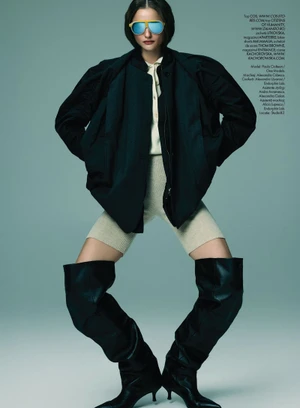 Model wearing blue mirrored sunglasses, dark jacket, cream top, knit shorts, and tall black boots against a plain grey backdrop.