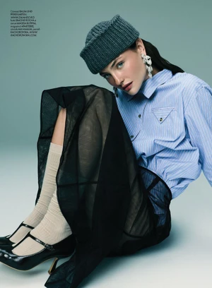 Model seated wearing a blue striped shirt, dark cap, sheer black skirt, cream socks, and black heels against a pale background.