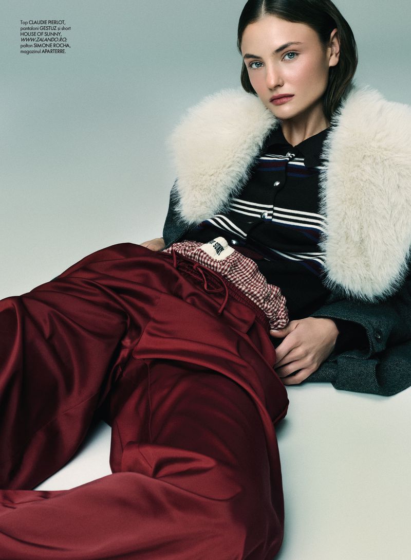 Model reclining wearing a dark striped polo shirt, white fur collar coat, and shiny red trousers on a white studio backdrop.