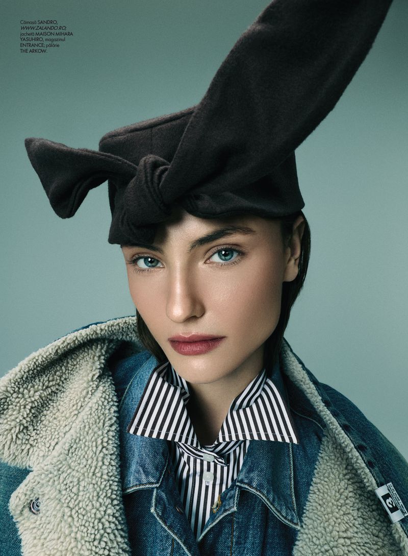 Model wearing a black knotted hat, striped shirt, and denim jacket with shearling lining against a teal background.