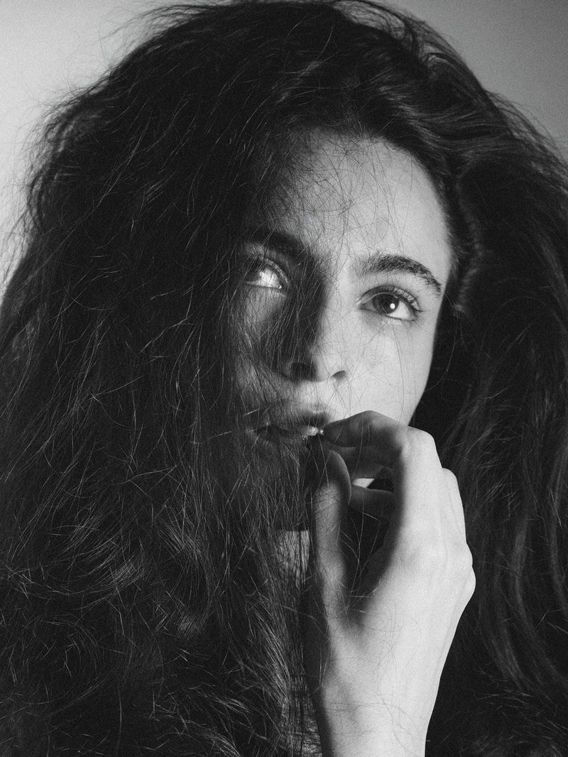Monochrome portrait of a woman with dark wavy hair gently holding strands near her lips against a neutral background.