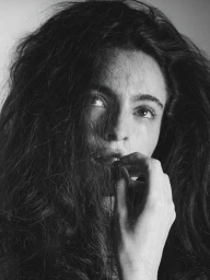 Monochrome portrait of a woman with dark wavy hair gently holding strands near her lips against a neutral background.