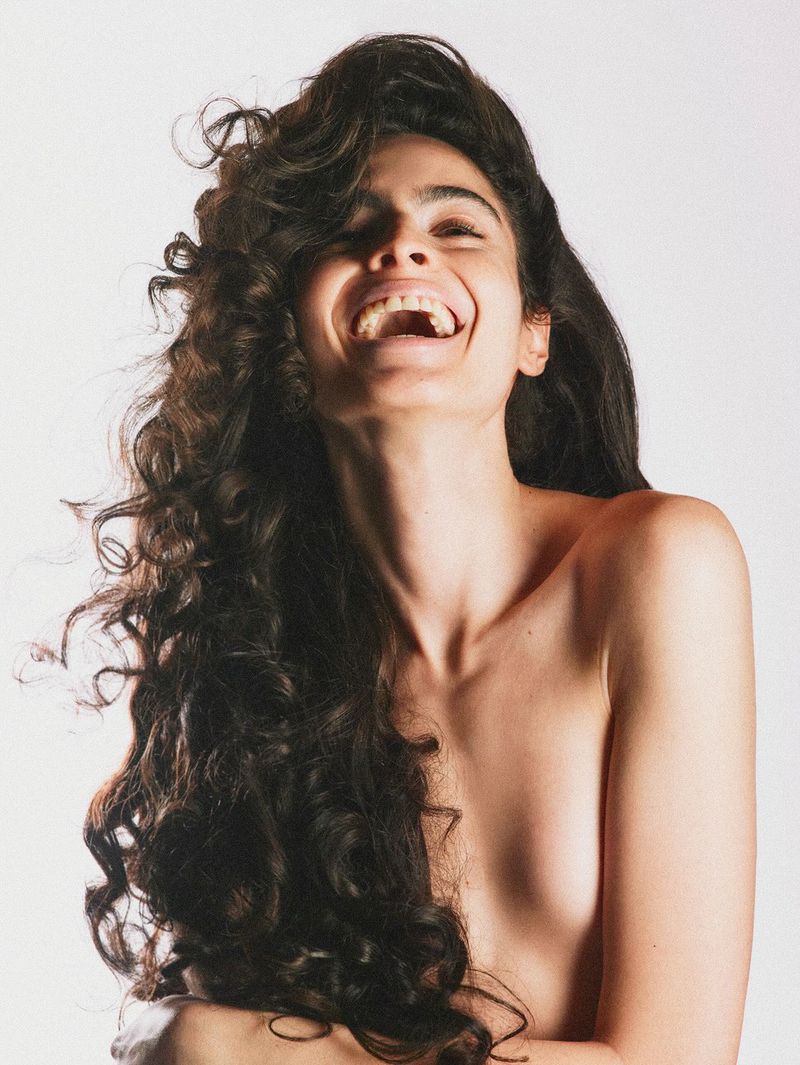 Close-up portrait of a laughing woman with long dark curly hair against a plain white background for i-D Magazine