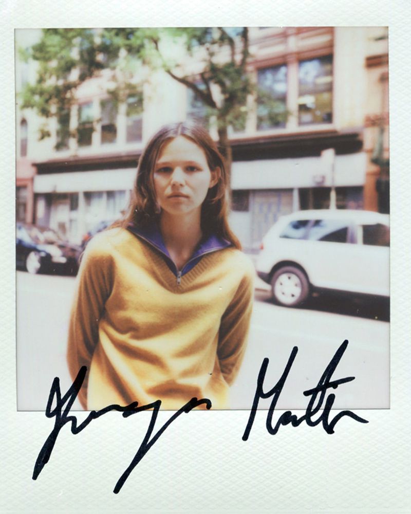 Polaroid photograph of a woman wearing a mustard yellow zip sweater standing on a city street with handwritten signature below.