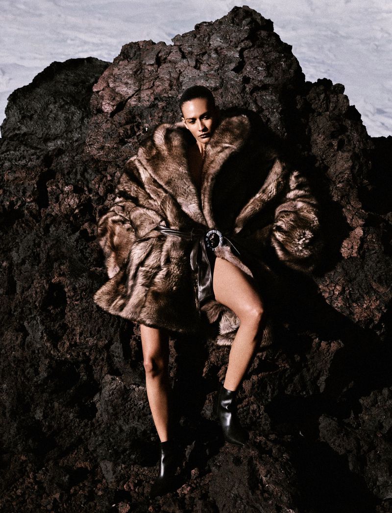 Model in a brown faux fur coat perched on dark volcanic rock for Numéro France