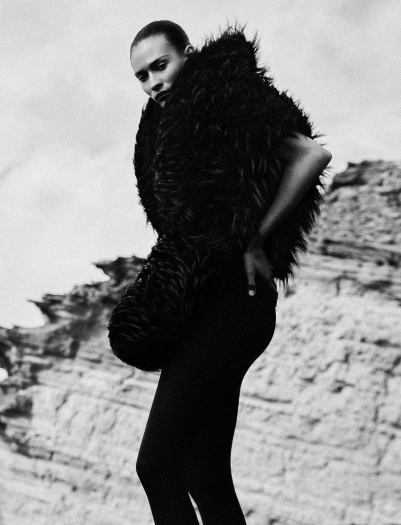 Model in voluminous black furry garment standing against a rocky cliff background in a black and white fashion portrait.