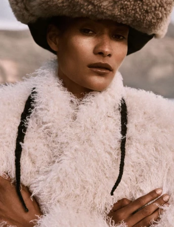 Model wearing a fluffy white textured coat and large fur hat with crossed arms in an outdoor setting.