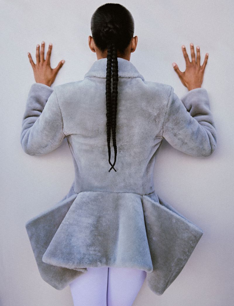 Model seen from behind wearing a grey peplum jacket with hands pressed against a light grey wall