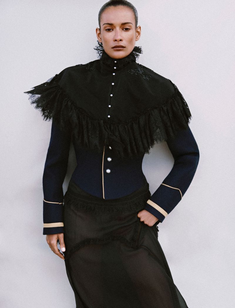 Model in a black structured high-neck jacket with ruffles and a sheer skirt against a plain white background for Numéro France