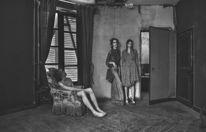 Black and white editorial fashion image featuring models posed inside a distressed room with worn walls and wooden parquet floors.