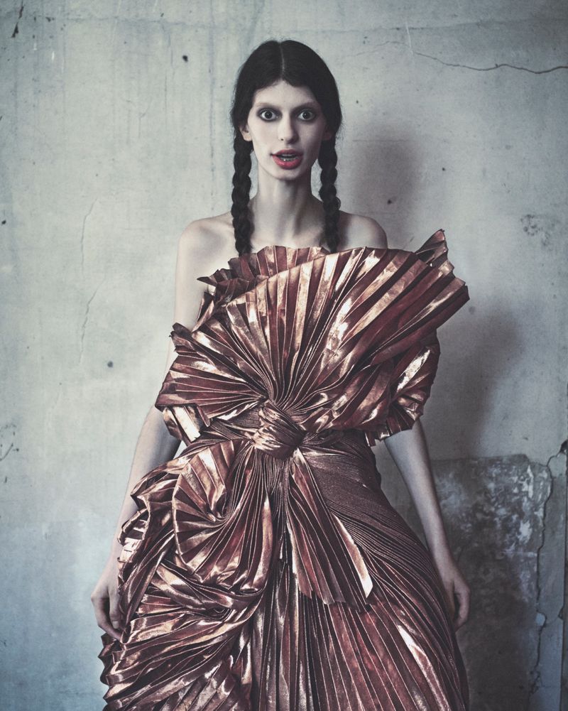 Model in a metallic pleated bronze dress with braided hair standing against a weathered concrete wall for Harper's Bazaar Italia.