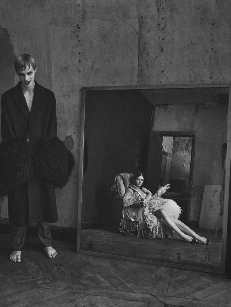 Monochrome fashion editorial featuring a man standing barefoot next to a large mirror reflecting a seated woman.