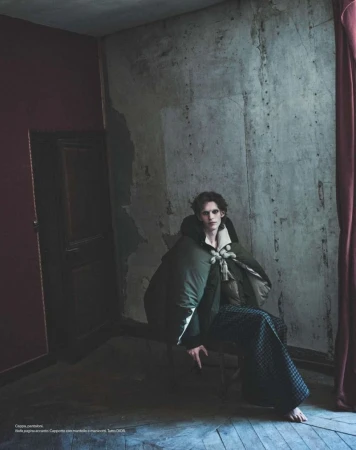 Model wearing a dark green coat and patterned trousers seated in a rustic room for Harper's Bazaar Italia
