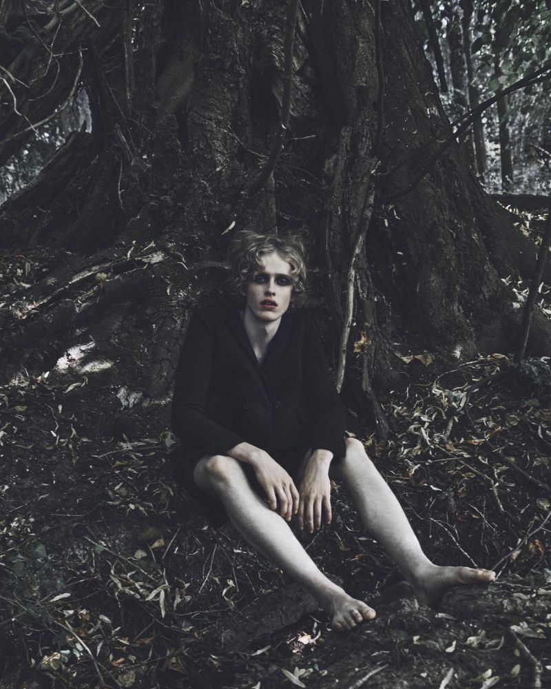 A young model in a dark jacket sitting at the base of a large tree in a forest for Harper's Bazaar Italia