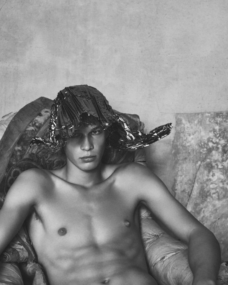 Monochrome portrait of a shirtless male model wearing a metallic foil hat seated on an ornate upholstered armchair.