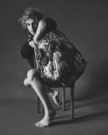 Monochrome portrait of a person with curly hair seated backward on a wooden chair, wearing a dark patterned garment.