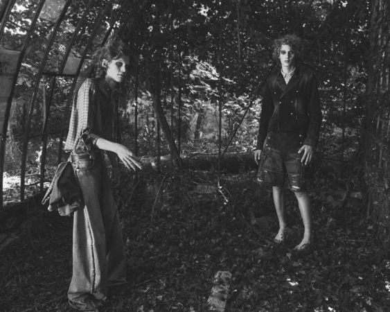 Two models standing inside a greenhouse structure overgrown with foliage in a black and white fashion editorial.