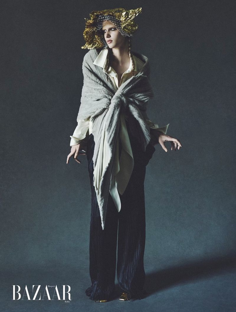 Model wearing a grey shawl, white blouse, and dark trousers with an ornate gold headpiece against a dark studio background