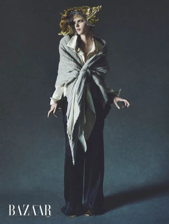 Model wearing a grey shawl, white blouse, and dark trousers with an ornate gold headpiece against a dark studio background