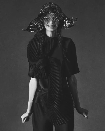 Model wearing a dark structured hat with netting and a pleated black top against a neutral studio background
