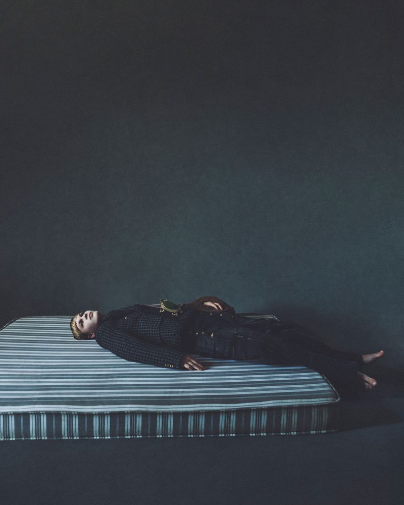 Model lying on a striped mattress against a dark textured background in an editorial fashion photograph.