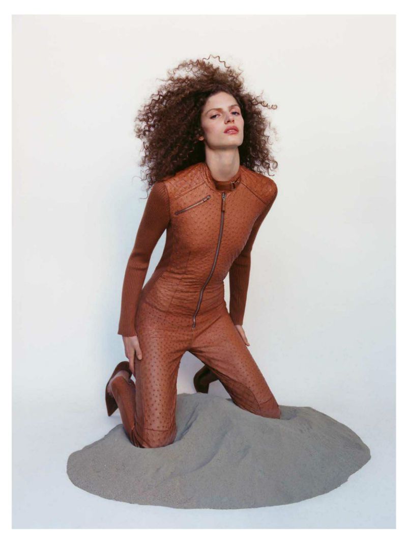 Model Sarah Isaksen kneeling in a sand pile wearing a fitted brown leather jumpsuit against a plain white background