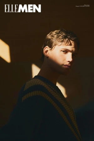 Close-up portrait of a young man wearing a dark sweater with stripes, lit by sharp sunlight against a plain dark wall.