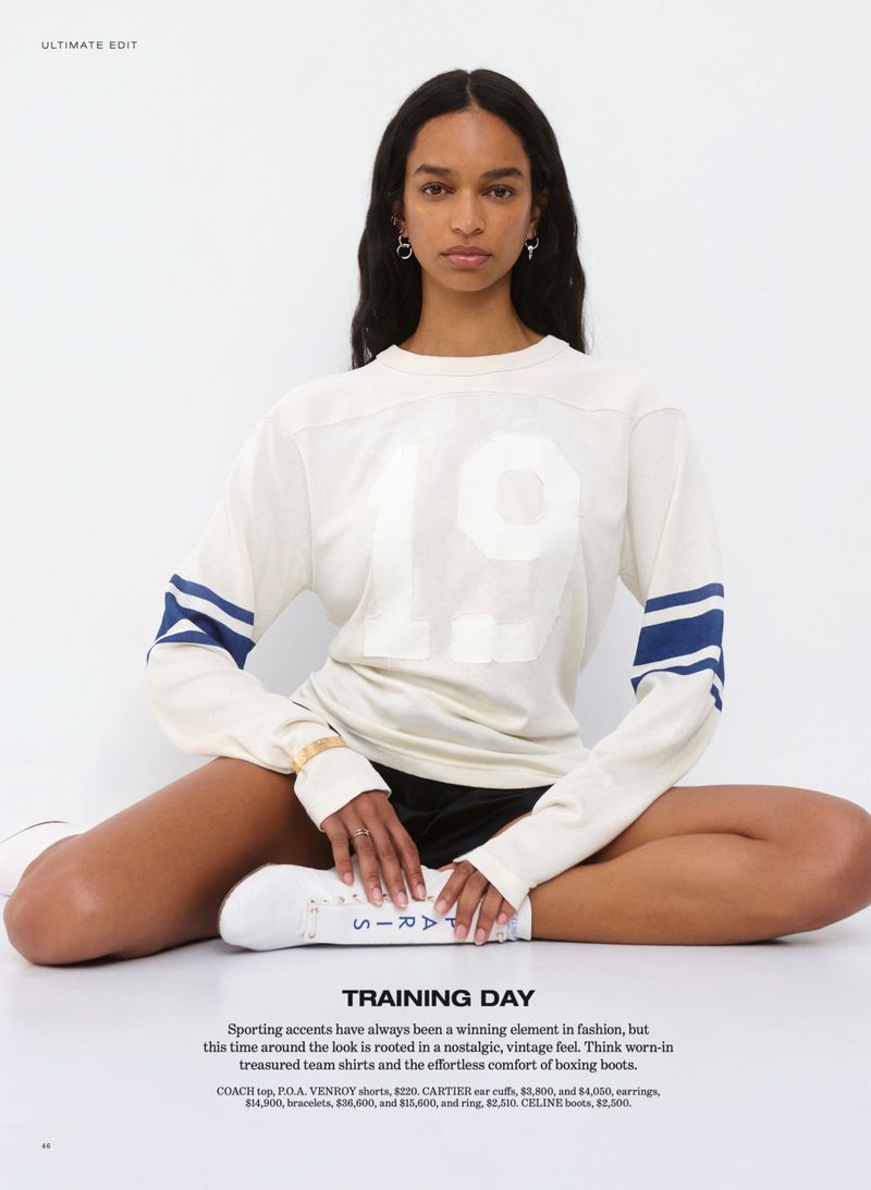 Model seated cross-legged wearing a cream sweater with blue sleeve stripes and dark shorts against a seamless white background.